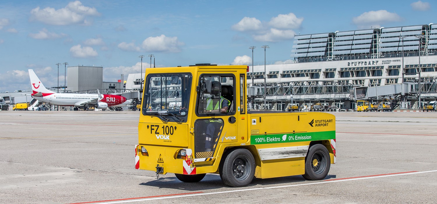 Ein elektrisch betriebener Gepäck-Schlepper steht auf dem Stuttgarter Flughafenvorfeld. Im Hintergrund stehen die Terminalgebäude. 