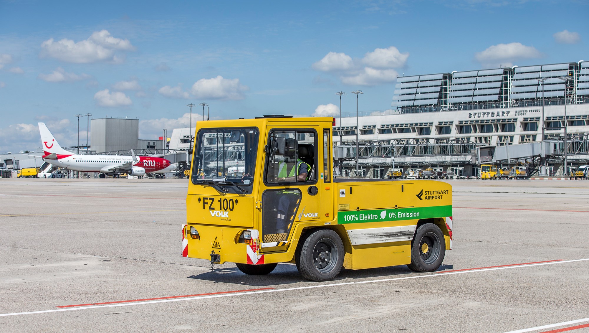 Ein elektrisch betriebener Gepäck-Schlepper steht auf dem Stuttgarter Flughafenvorfeld. Im Hintergrund stehen die Terminalgebäude. 