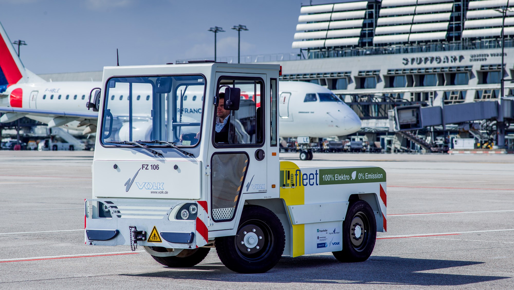 Ein elektrisch betriebener Gepäck-Schlepper mit Litium-Ionen-Batterie steht auf dem Stuttgarter Flughafenvorfeld. Im Hintergrund rollt ein Flugzeug zur Startposition. 