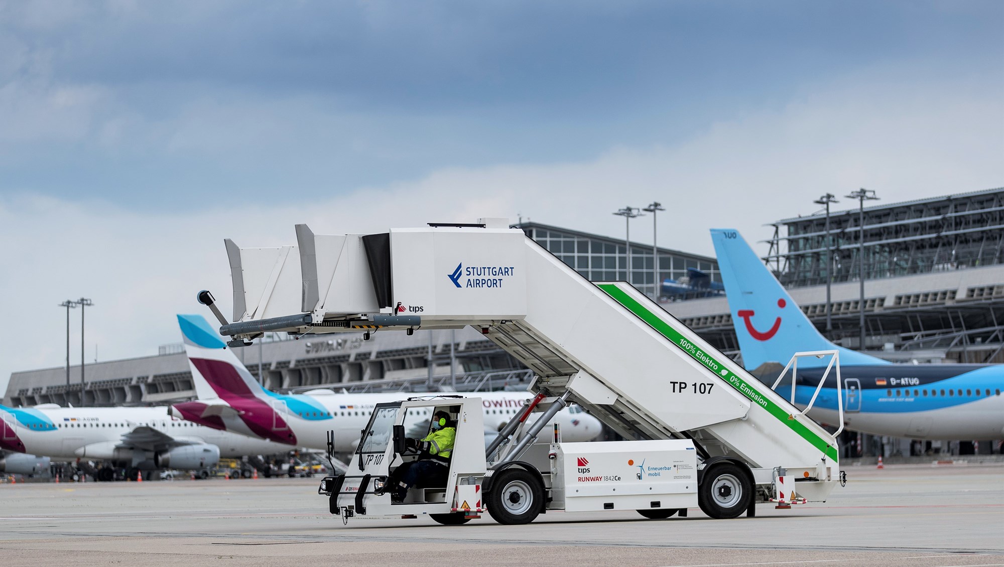 Eine elektrisch betriebene Passagiertreppe steht auf dem Stuttgarter Flughafenvorfeld. Im Hintergrund parken mehrere Flugzeuge. 